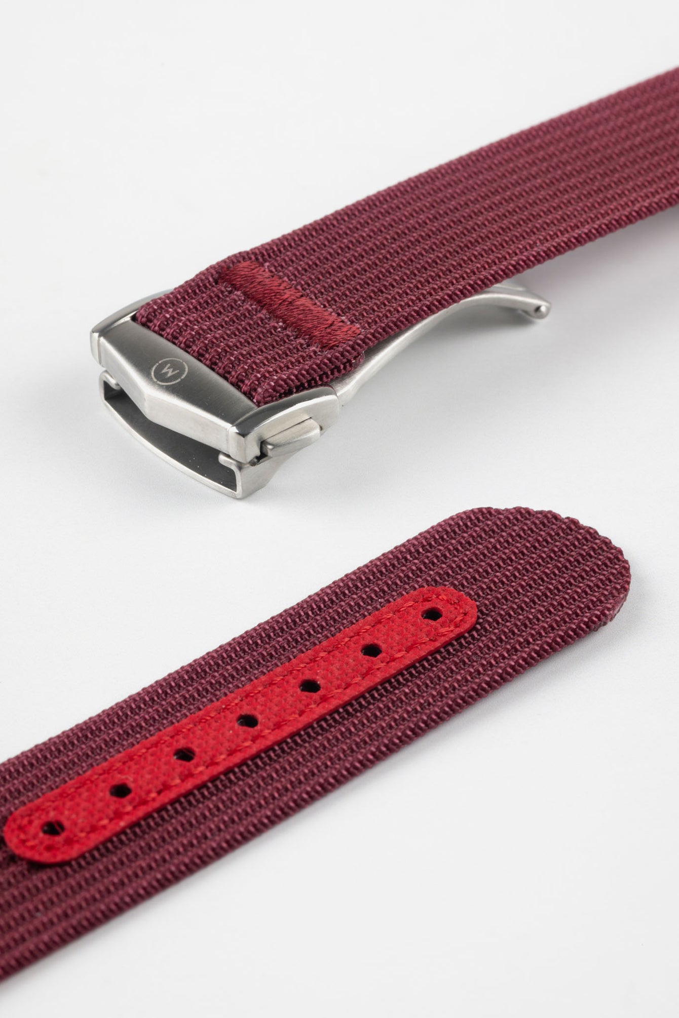 A three-quarter view of a burgundy ribbed-nylon watch strap fastened into a loop, highlighting the brushed metal buckle and woven texture on a white background.