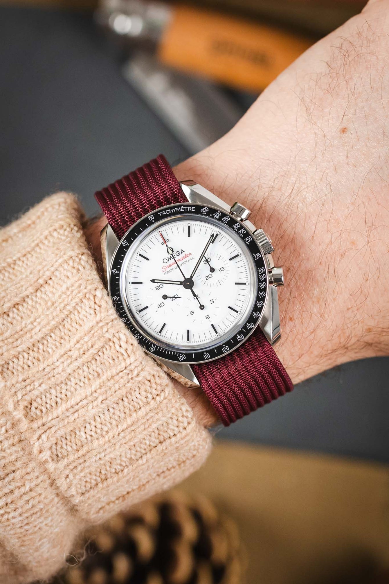 A close-up shot of a person's wrist wearing a Omega Speedmaster with a white dial. The watch is paired with a burgundy ribbed-nylon strap and is worn over the sleeve of a beige cable-knit sweater, creating a cozy, outdoor-inspired look.