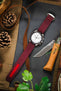 A top-down lifestyle shot of a Omega Speedmaster watch with a white dial, fitted on a burgundy ribbed-nylon strap. The watch rests on a rustic leather pouch atop a wooden surface, surrounded by pine cones, an Opinel folding knife, and green foliage.