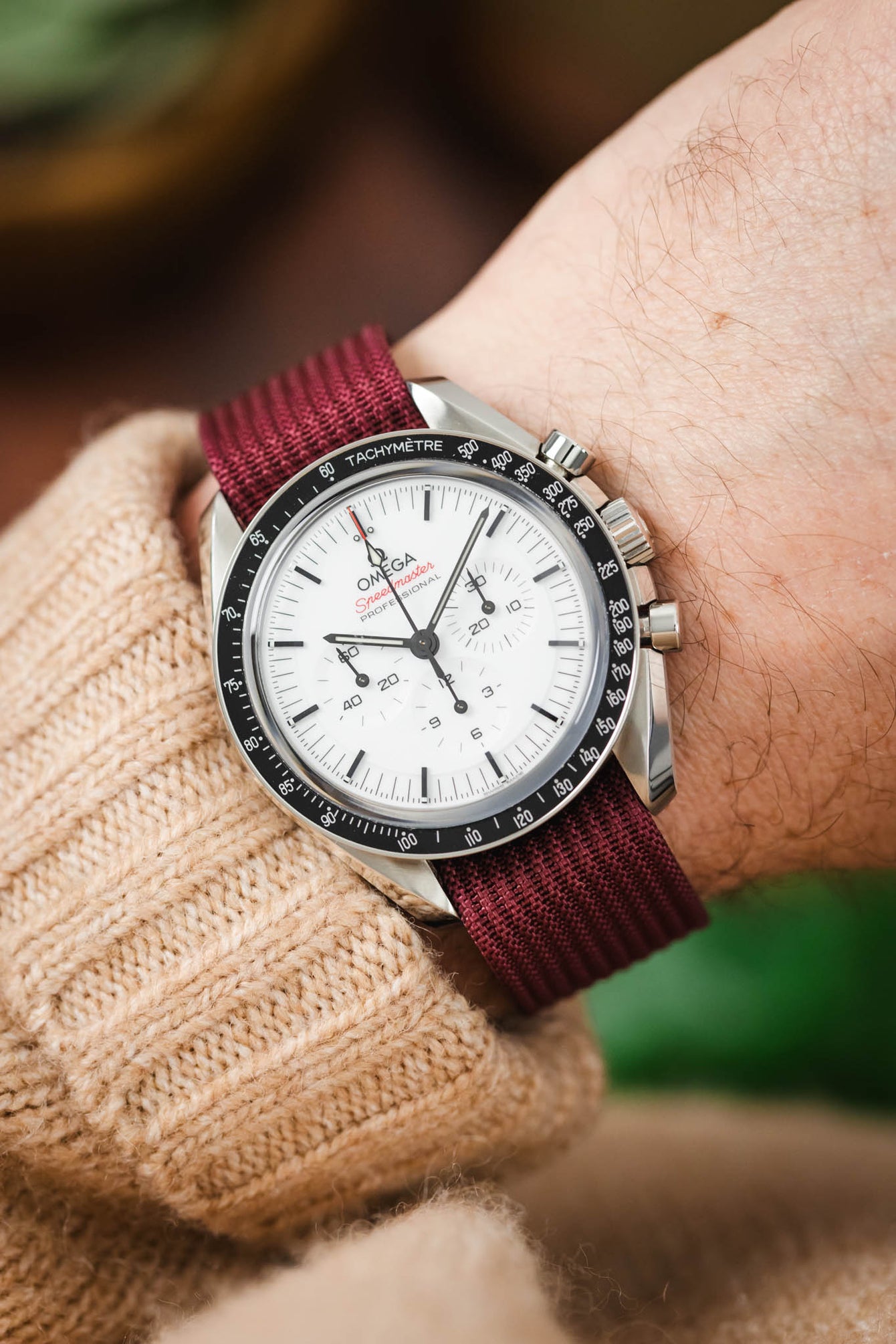 Close-up of a stainless steel Omega Speedmaster chronograph with a white dial and black tachymeter bezel, fitted on a burgundy fabric strap, resting on a wooden surface with a book, a metal tool, and softly blurred greenery in the foreground.