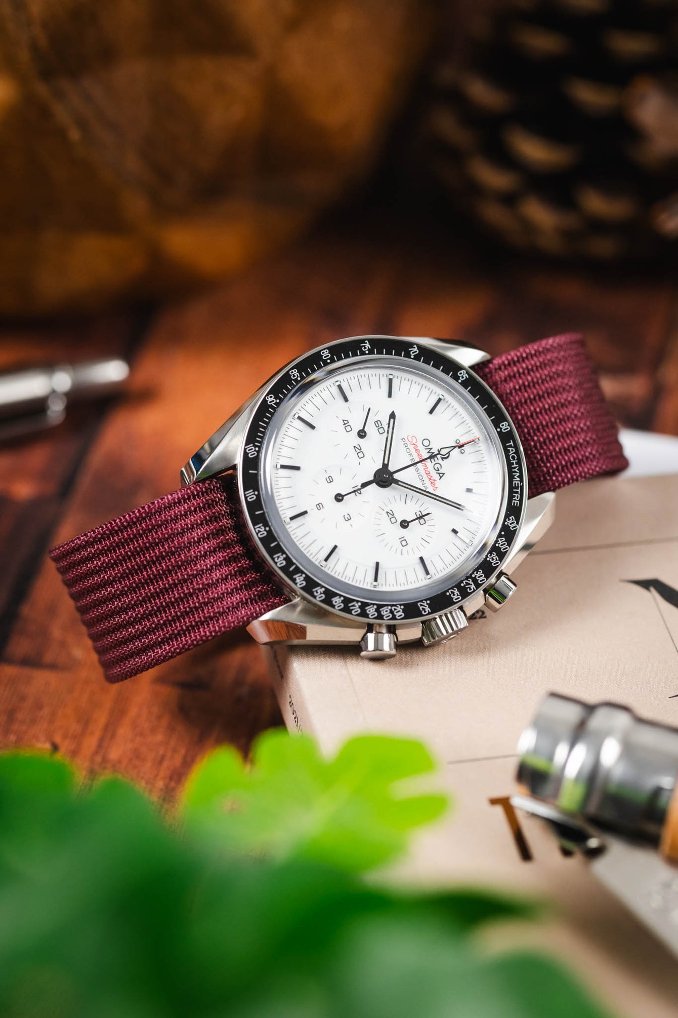 Close-up of a stainless steel Omega Speedmaster chronograph with a white dial and black tachymeter bezel, fitted on a burgundy fabric strap, resting on a wooden surface with a book, a metal tool, and softly blurred greenery in the foreground.