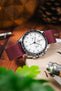 Close-up of a stainless steel Omega Speedmaster chronograph with a white dial and black tachymeter bezel, fitted on a burgundy fabric strap, resting on a wooden surface with a book, a metal tool, and softly blurred greenery in the foreground.