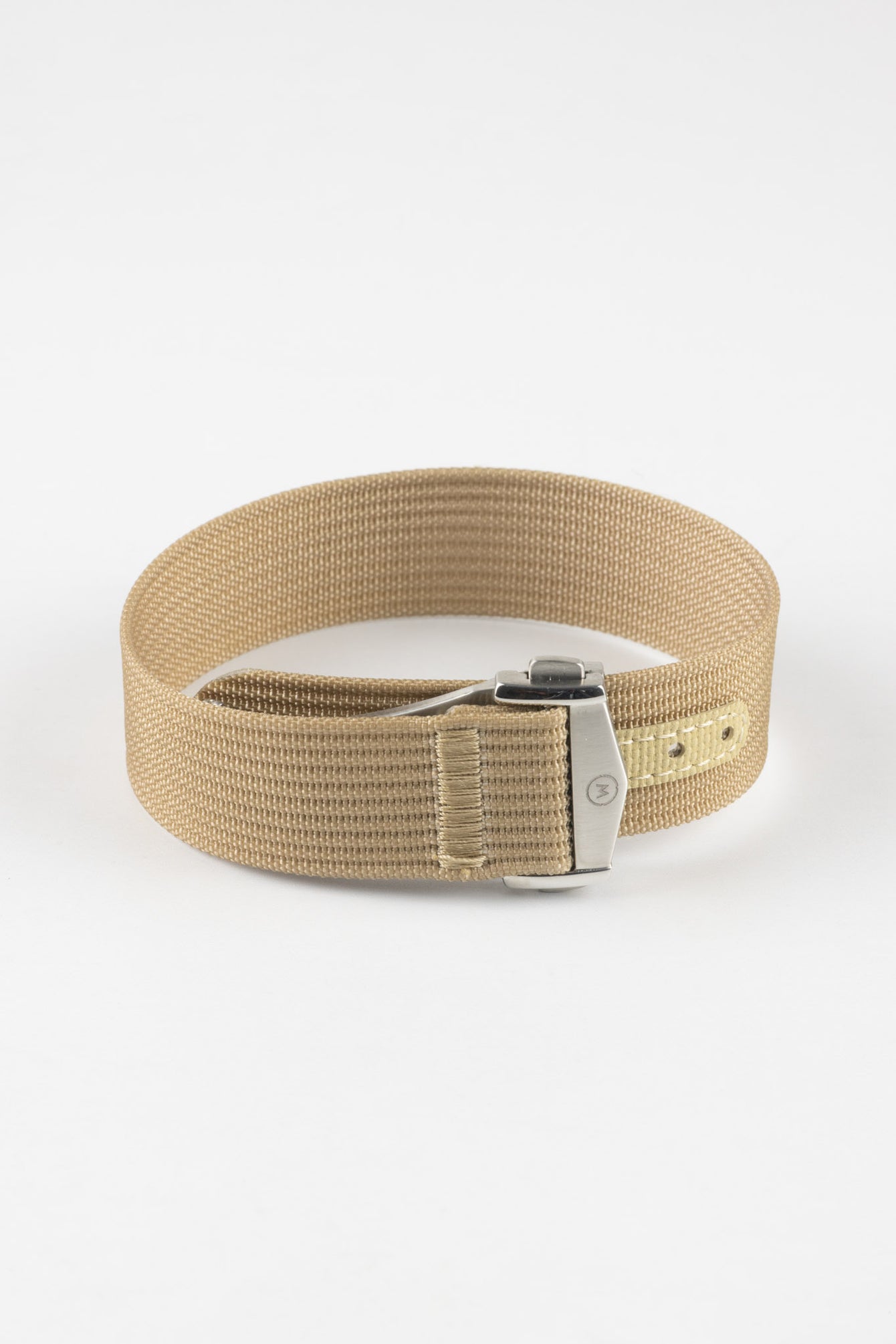 
Close-up of a light cream ribbed nylon watch strap arranged in a loop on a light grey background. The strap features a textured woven finish and a brushed silver deployant clasp with an engraved logo. A matching cream reinforced section with evenly spaced adjustment holes is visible near the clasp, highlighting the strap’s construction and minimalist design.