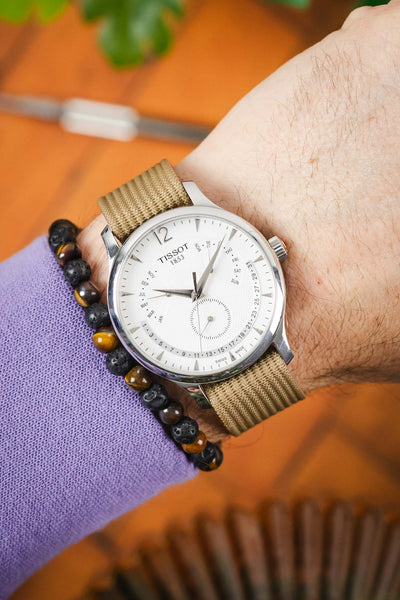 Close-up of a Tissot watch with a white multi-dial face on a beige nylon strap, worn on a wrist next to a beaded bracelet and lavender sleeve, with a warm blurred background.