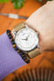 Close-up of a Tissot watch with a white multi-dial face on a beige nylon strap, worn on a wrist next to a beaded bracelet and lavender sleeve, with a warm blurred background.