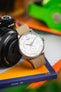 Close-up of a Tissot watch with a white multi-dial face and beige nylon strap, resting on books beside a camera lens, with a colorful blurred background.