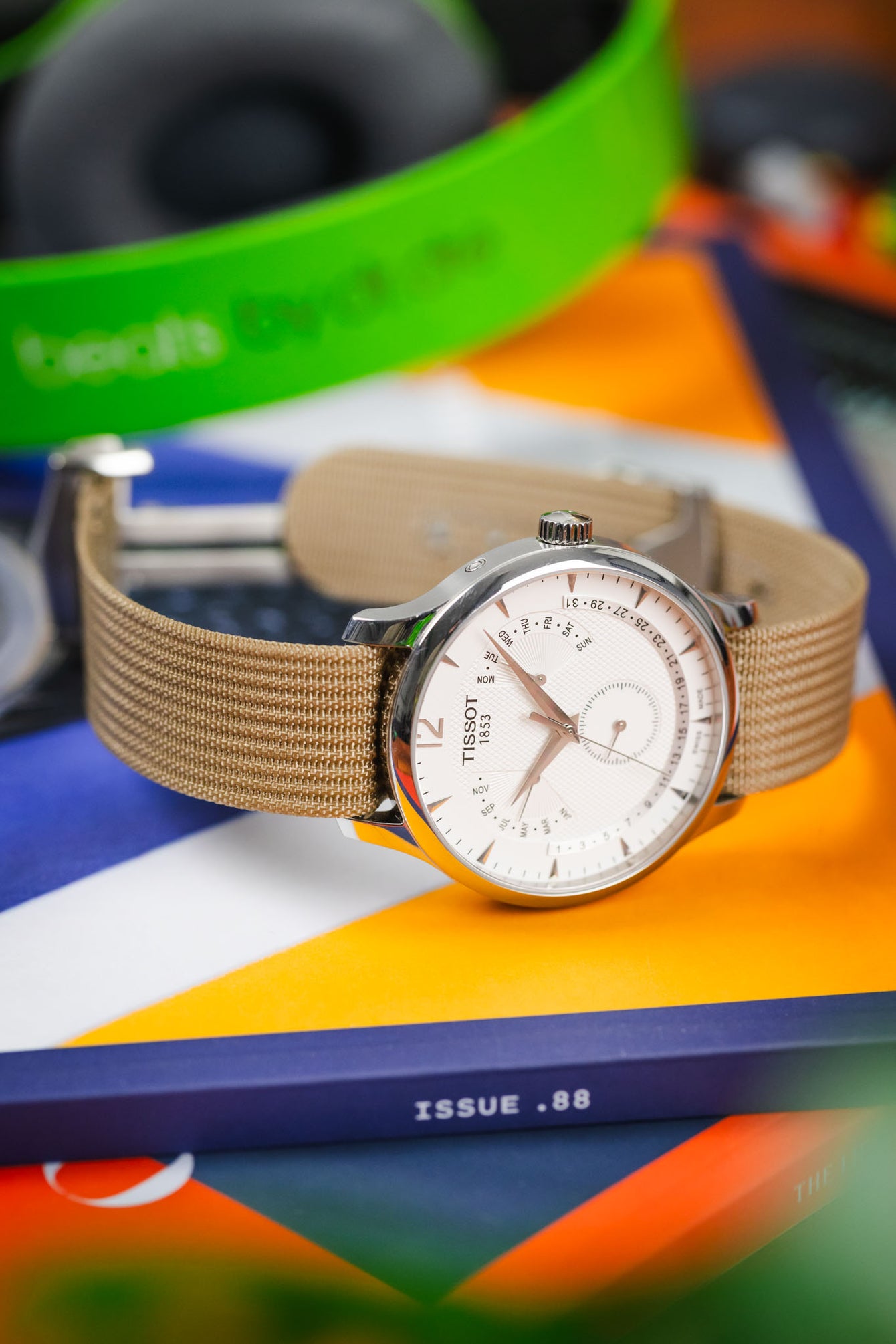Close-up of a Tissot watch with a white multi-dial face and beige nylon strap resting on books, with a blurred colorful background and nearby headphones.