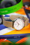 Close-up of a Tissot watch with a white multi-dial face and beige nylon strap resting on books, with a blurred colorful background and nearby headphones.