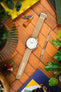 Flat-lay of a Tissot watch with a white multi-dial face and beige nylon strap on a wooden surface, surrounded by everyday items like earbuds, a small knife, tools, plants, and books in a colorful arrangement.