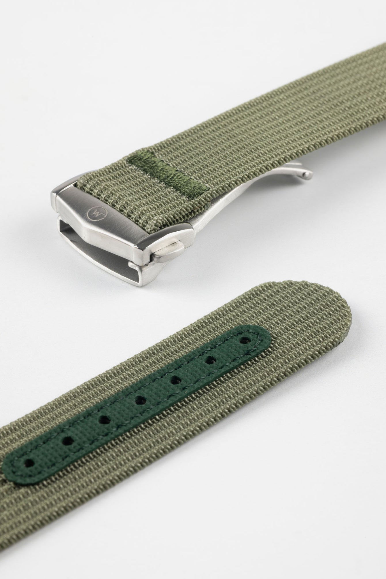 A close-up, angled shot of a green ribbed-nylon watch strap showing both the brushed stainless steel deployment buckle and the adjustment holes. The image highlights the fine woven texture of the fabric and the precision stitching near the metal clasp.