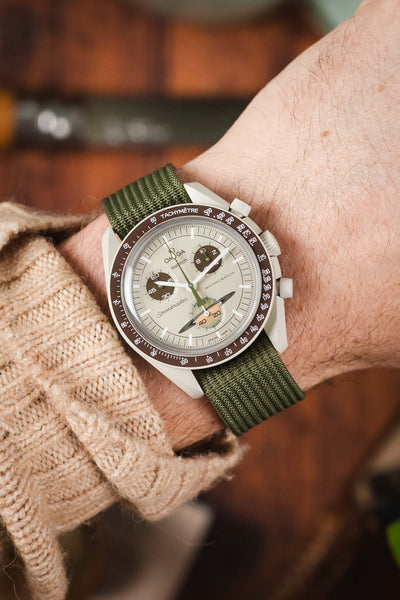 A close-up shot of a person's wrist wearing a Omega X Swatch MoonSwatch Saturn watch with a "saturn" dial. The watch is paired with a green ribbed-nylon strap and is worn over the sleeve of a beige cable-knit sweater, creating a cozy, outdoor-inspired look.