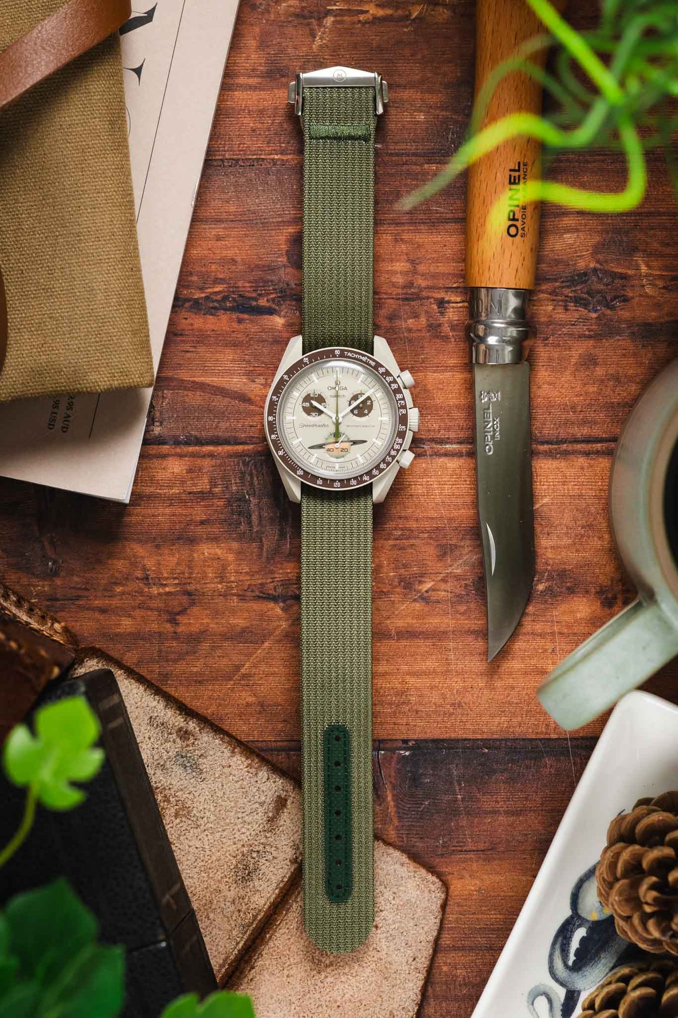 A top-down lifestyle shot of a Omega X Swatch MoonSwatch Saturn chronograph watch with a beige, fitted on a green ribbed-nylon strap. The watch rests on a rustic leather pouch atop a wooden surface, surrounded by pine cones, an Opinel folding knife, and green foliage.