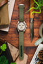 A top-down lifestyle shot of a Omega X Swatch MoonSwatch Saturn chronograph watch with a beige, fitted on a green ribbed-nylon strap. The watch rests on a rustic leather pouch atop a wooden surface, surrounded by pine cones, an Opinel folding knife, and green foliage.