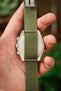 Close-up of a green ribbed nylon watch strap fitted to a watch case, shown from the back and held in a hand, with metal spring bars visible and a blurred natural background.