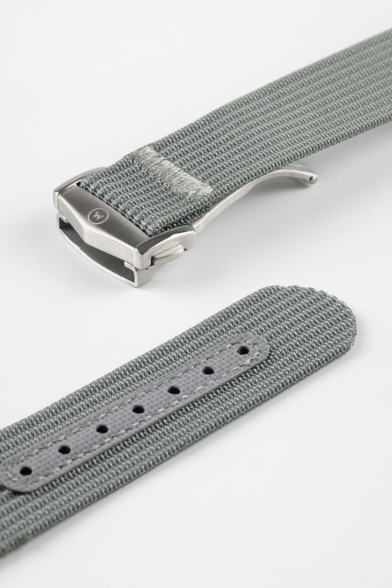 A three-quarter view of a grey ribbed-nylon watch strap fastened into a loop, highlighting the brushed metal buckle and woven texture on a white background.