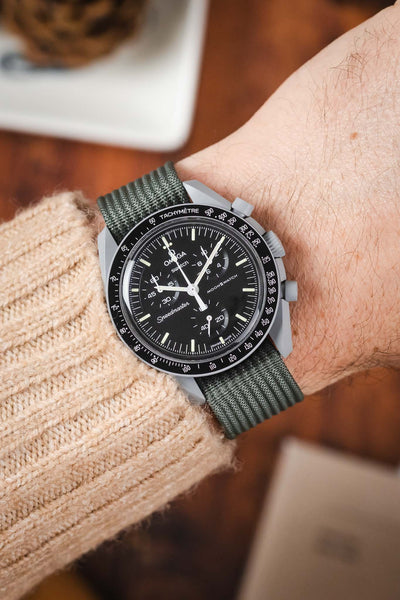 A close-up shot of a person's wrist wearing a Omega X Swatch MoonSwatch Moon. The watch is paired with a grey ribbed-nylon strap and is worn over the sleeve of a beige cable-knit sweater, creating a cozy, outdoor-inspired look.