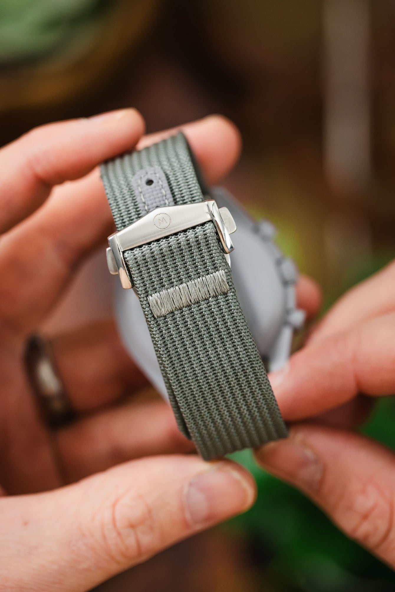 Close-up of a grey ribbed nylon watch strap held in hand, showing a brushed silver clasp with an engraved logo and a reinforced stitched section near the buckle, against a blurred background.