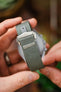 Close-up of a grey ribbed nylon watch strap held in hand, showing a brushed silver clasp with an engraved logo and a reinforced stitched section near the buckle, against a blurred background.