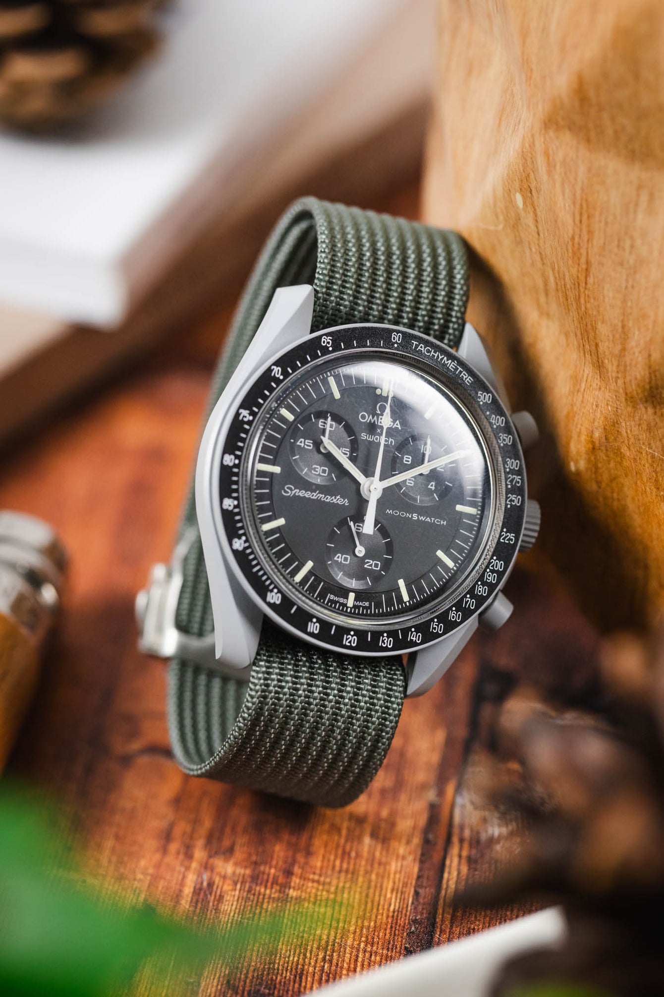 Chronograph-style Omega watch with a black dial and tachymeter bezel on a green ribbed nylon strap, resting on a wooden surface with blurred lifestyle elements in the background.