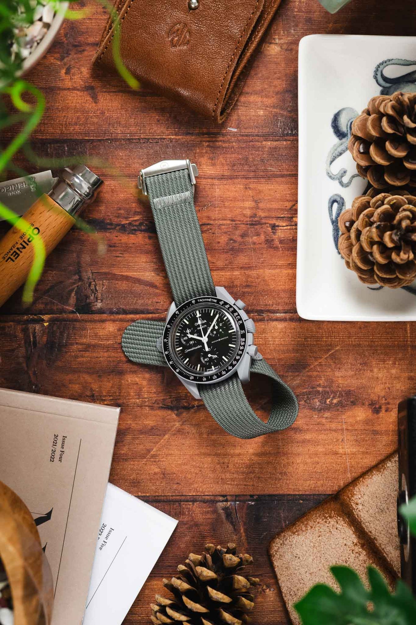 A top-down lifestyle shot of a Omega X Swatch MoonSwatch Moon watch with a black dial, fitted on a grey ribbed-nylon strap. The watch rests on a rustic leather pouch atop a wooden surface, surrounded by pine cones, an Opinel folding knife, and green foliage.