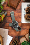 A top-down lifestyle shot of a Omega X Swatch MoonSwatch Moon watch with a black dial, fitted on a grey ribbed-nylon strap. The watch rests on a rustic leather pouch atop a wooden surface, surrounded by pine cones, an Opinel folding knife, and green foliage.