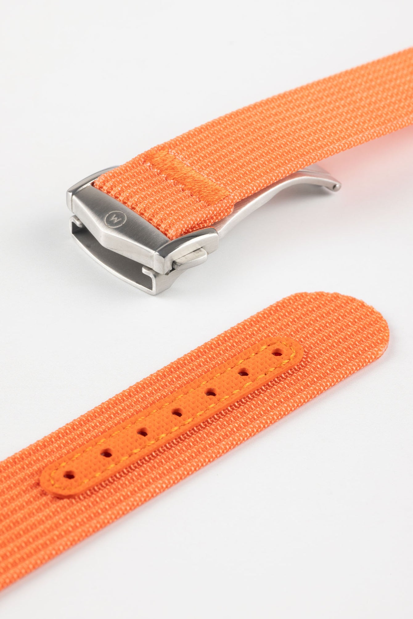 Angled close-up of a bright orange ribbed nylon watch strap highlighting the textured woven surface and a brushed silver deployant clasp with an engraved logo. A separate strap section shows an orange reinforced panel with neatly stitched edges and evenly spaced adjustment holes, all displayed against a clean light grey background.