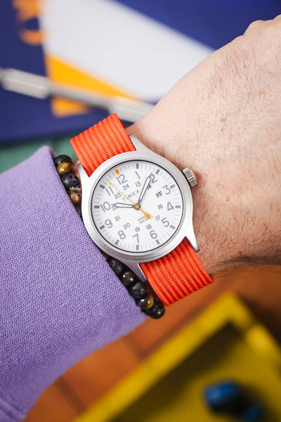 Close-up of a wrist wearing a Timex analog watch with a white dial, gray case, and a bright orange ribbed nylon strap. The dial features large gray Arabic numerals, a 24-hour inner scale, and an orange seconds hand, with “TIMEX” and “INDIGLO WR 50M” visible. A beaded bracelet with dark and brown stones sits beside the watch, partially covered by a lavender sleeve. The background is softly blurred with blue, yellow, and orange tones, creating a colorful, vibrant setting.