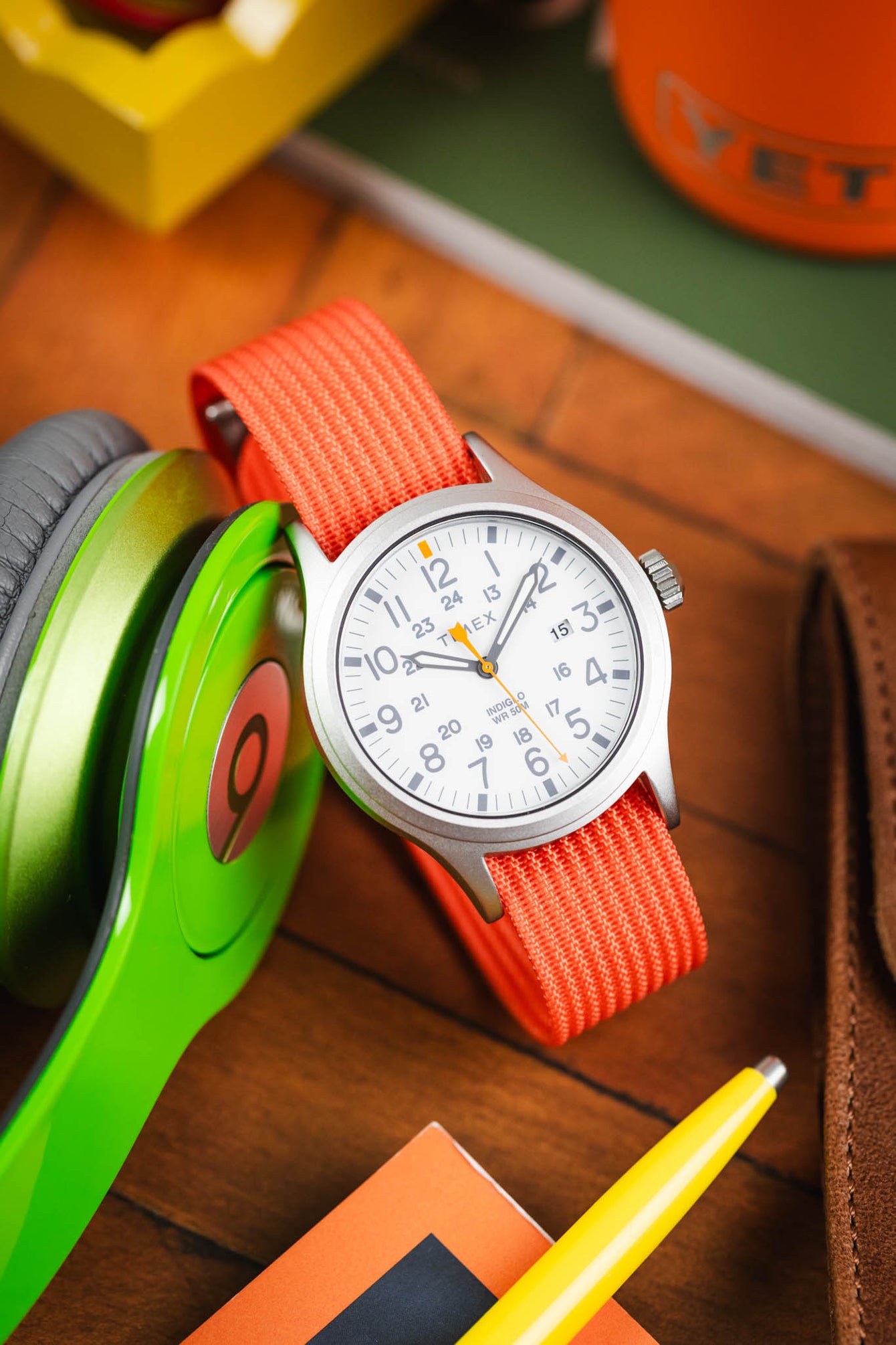 Close-up product shot of a Timex analog wristwatch with a white dial and matte gray case, fitted with a bright orange ribbed nylon strap. The dial features large gray Arabic numerals, an inner 24‑hour scale, and an orange seconds hand, with “TIMEX” and “INDIGLO WR 50M” visible. The watch is propped at an angle on a wooden surface beside green over‑ear headphones, with surrounding items including a brown leather wallet, a yellow pen, and colorful objects in soft focus, creating a vibrant, warm-toned scene.