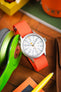Close-up product shot of a Timex analog wristwatch with a white dial and matte gray case, fitted with a bright orange ribbed nylon strap. The dial features large gray Arabic numerals, an inner 24‑hour scale, and an orange seconds hand, with “TIMEX” and “INDIGLO WR 50M” visible. The watch is propped at an angle on a wooden surface beside green over‑ear headphones, with surrounding items including a brown leather wallet, a yellow pen, and colorful objects in soft focus, creating a vibrant, warm-toned scene.