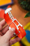Close-up of an orange nylon watch strap with an open metal clasp, held in hand, showing its textured weave, hinges, and adjustment holes against a blurred, colorful background.
