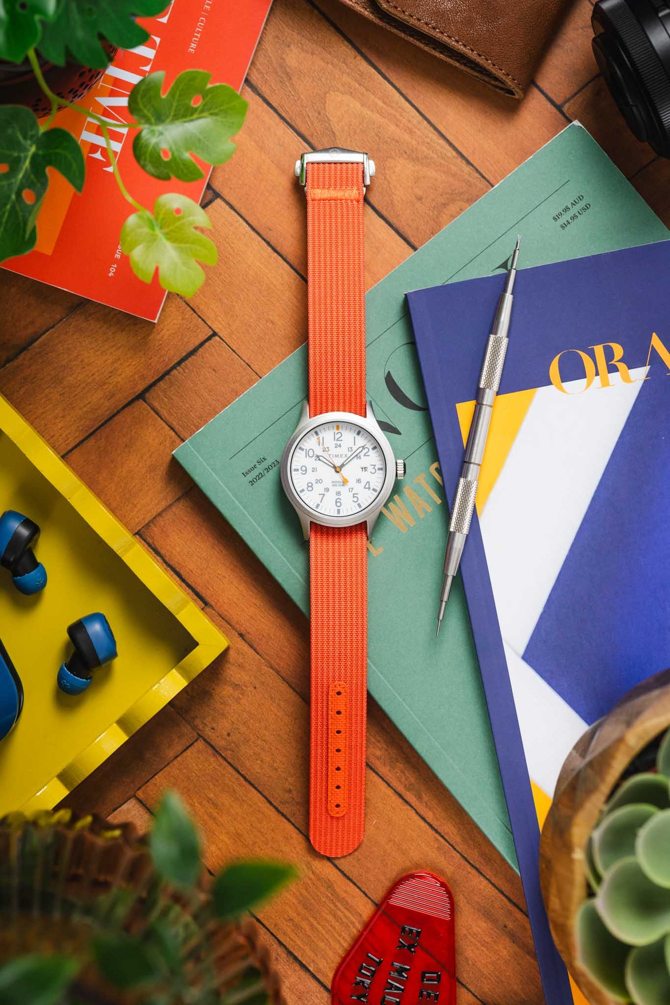 Flat-lay photo of a wristwatch with a bright orange ribbed nylon strap and a white dial, placed vertically on books and magazines on a wooden floor. Surrounding items include a metal precision tool, a brown leather wallet, a small potted plant, wireless earbuds in a yellow tray, and a red object with the word “EXIT.” The scene features vivid colors and soft natural lighting.