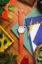 Flat-lay photo of a wristwatch with a bright orange ribbed nylon strap and a white dial, placed vertically on books and magazines on a wooden floor. Surrounding items include a metal precision tool, a brown leather wallet, a small potted plant, wireless earbuds in a yellow tray, and a red object with the word “EXIT.” The scene features vivid colors and soft natural lighting.