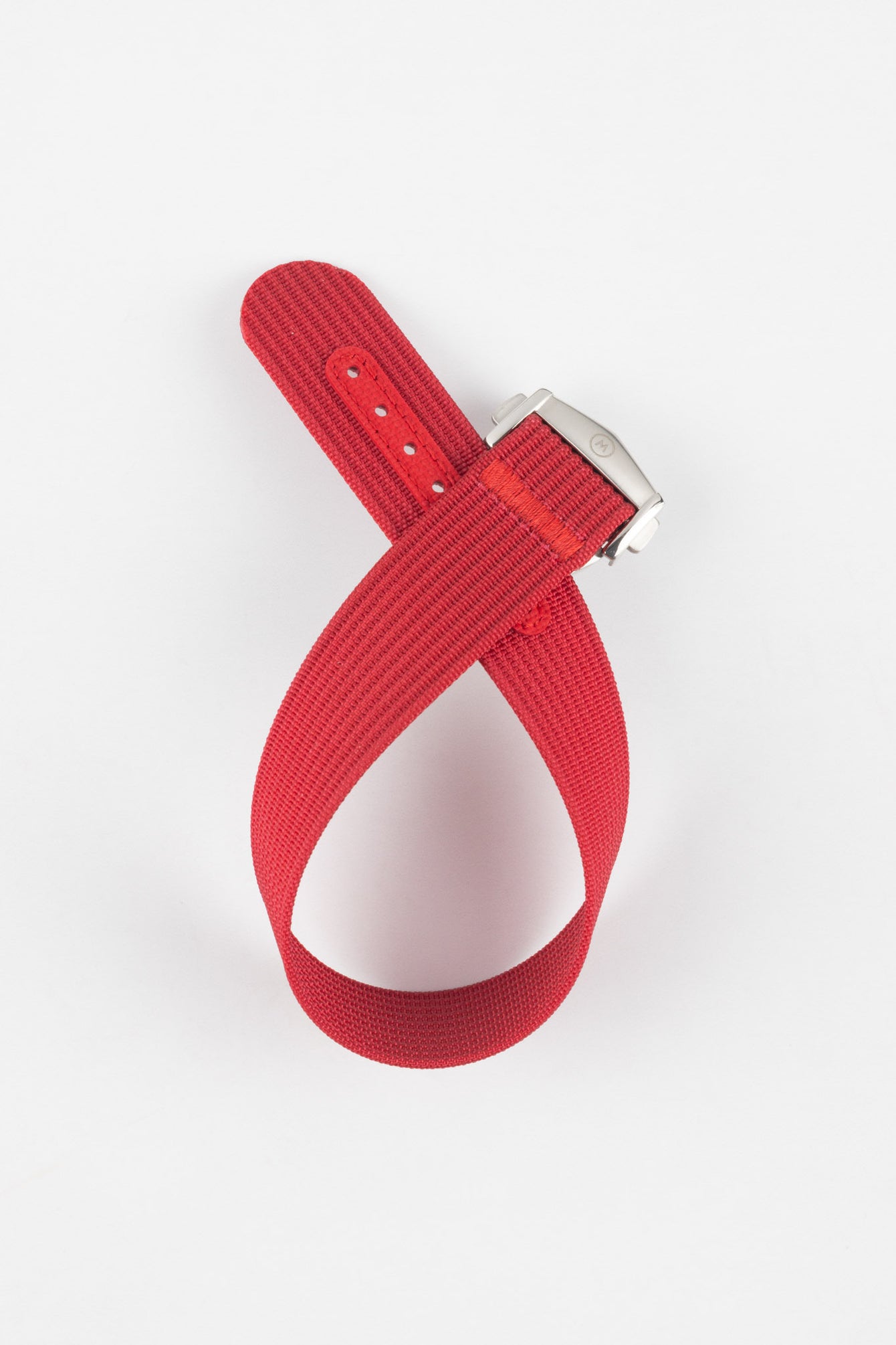 Red ribbed nylon watch strap displayed looped on a light grey background, featuring a textured woven surface and a brushed silver deployant clasp with an engraved logo. The strap includes a matching red reinforced section with evenly spaced adjustment holes, highlighting its bold color and durable construction.