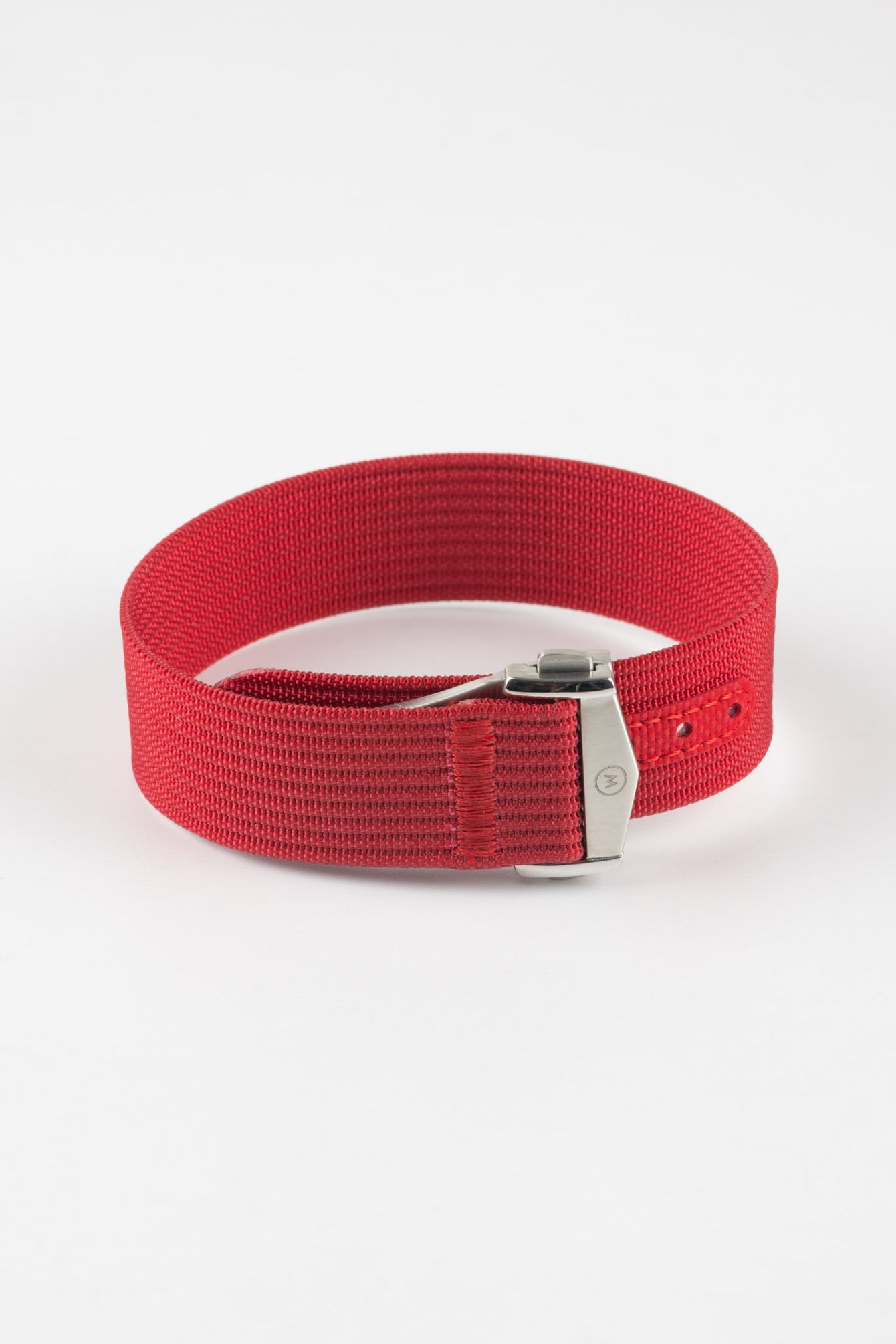 Close-up of a red ribbed nylon watch strap arranged in a loop on a light grey background. The strap features a textured woven finish and a brushed silver deployant clasp with an engraved logo. A red reinforced section with evenly spaced adjustment holes is visible near the clasp, highlighting the strap’s construction and bold color.