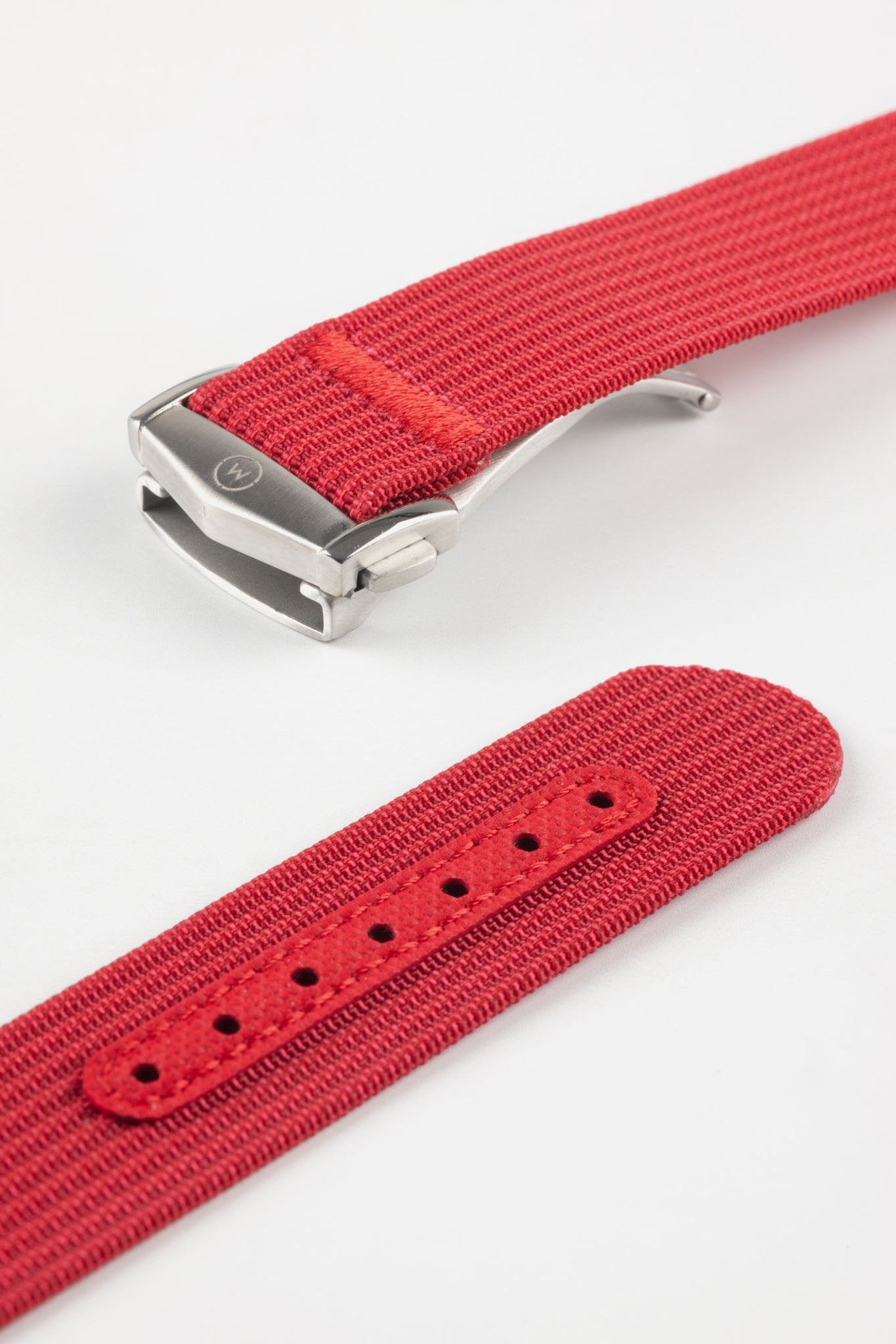 Angled close-up of a red ribbed nylon watch strap showing the textured woven surface and a brushed silver deployant clasp with an engraved logo. A separate strap section displays a matching red reinforced panel with neatly stitched edges and evenly spaced adjustment holes, all set against a clean light grey background.