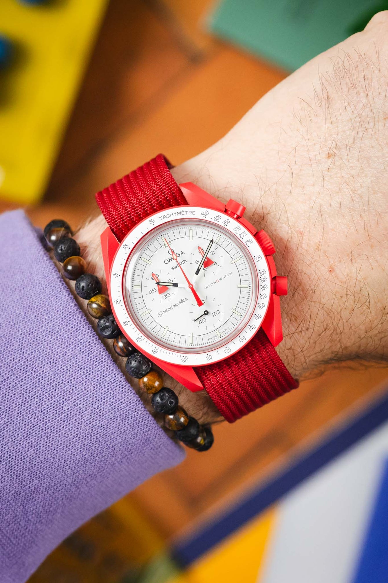 Close-up of a red Omega × Swatch Speedmaster “MoonSwatch” wristwatch with a white chronograph dial and red fabric strap, worn on a wrist alongside a beaded bracelet, against a colorful blurred background.