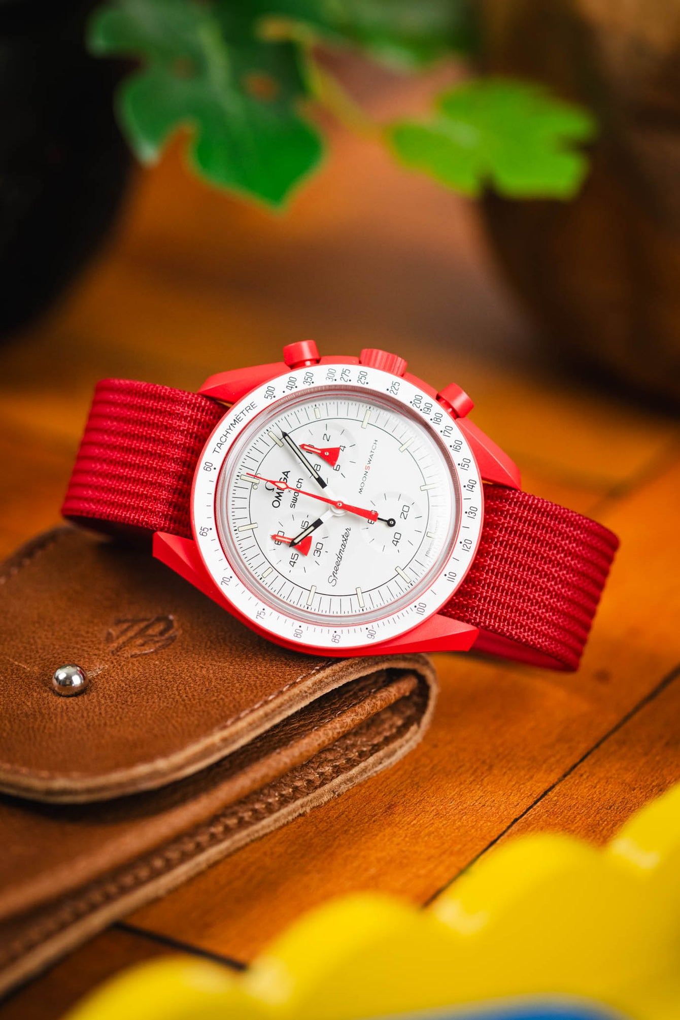 Close-up product shot of a red Omega × Swatch Speedmaster “MoonSwatch” wristwatch with a white chronograph dial and red tachymeter bezel, paired with a red textured nylon strap. The watch is placed on a brown leather wallet on a wooden surface. The background is softly blurred with warm orange tones and green leaves, creating a vibrant, colorful backdrop.