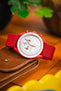 Close-up product shot of a red Omega × Swatch Speedmaster “MoonSwatch” wristwatch with a white chronograph dial and red tachymeter bezel, paired with a red textured nylon strap. The watch is placed on a brown leather wallet on a wooden surface. The background is softly blurred with warm orange tones and green leaves, creating a vibrant, colorful backdrop.
