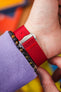 Close-up of the underside of a wrist wearing a bright red textured nylon watch strap with a polished metal buckle engraved with an “M” logo. The strap is being fastened, with adjustment holes visible. A beaded bracelet with dark and brown stones sits next to the strap, and a lavender sleeve partially covers the wrist. The background is softly blurred with warm orange and multicolored tones.