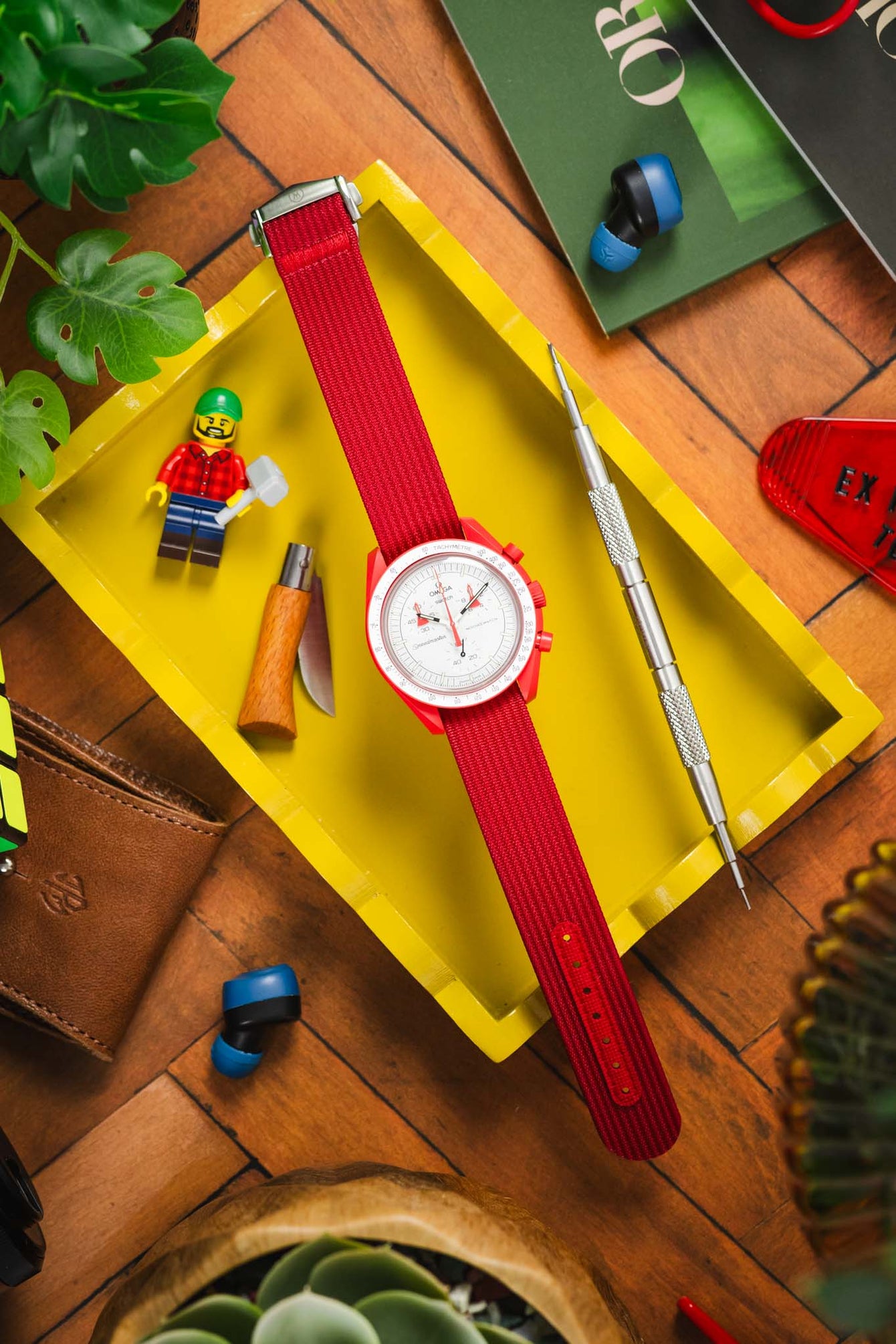 Flat lay of a white-dial wristwatch with a vivid red ribbed fabric strap placed inside a yellow tray, surrounded by assorted items including a small toy figure, a wooden-handled tool, a precision screwdriver, blue wireless earbuds, a green book, and leafy plants on a wooden surface.