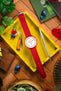 Flat lay of a white-dial wristwatch with a vivid red ribbed fabric strap placed inside a yellow tray, surrounded by assorted items including a small toy figure, a wooden-handled tool, a precision screwdriver, blue wireless earbuds, a green book, and leafy plants on a wooden surface.