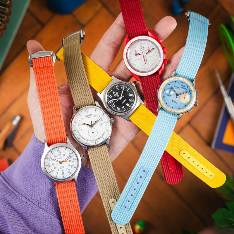 A hand holding a collection of wristwatches with vibrant ribbed nylon straps in orange, beige, red, yellow, and light blue, each featuring distinct dial designs including simple analog faces and a chronograph. The watches are fanned out across the palm, showcasing different case styles and strap textures. The background is softly blurred with warm wooden flooring and colorful objects, emphasizing the bright, summery tones and variety of the straps.