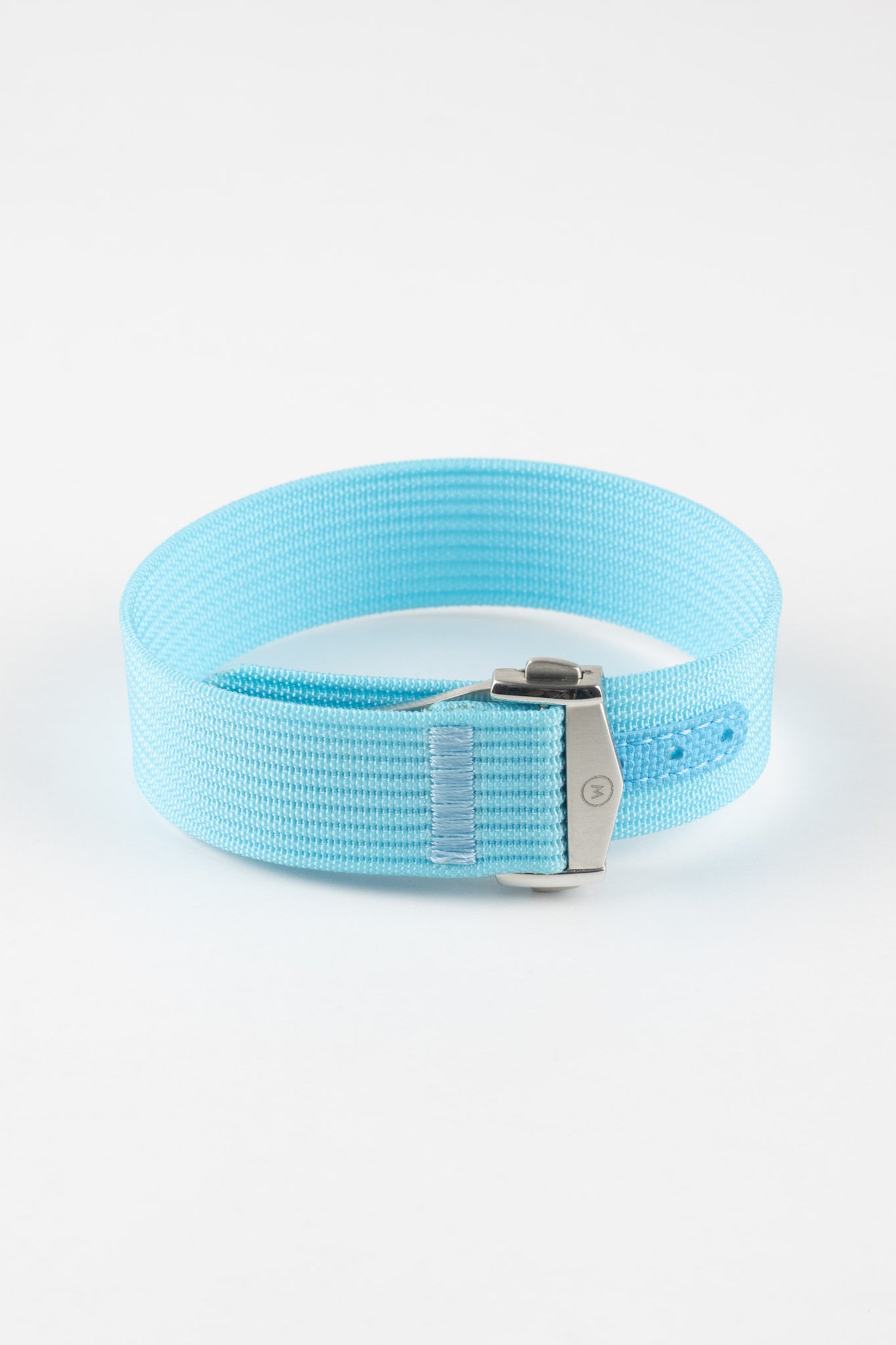 
Close-up of a light turquoise ribbed nylon watch strap arranged in a loop on a light grey background. The strap features a fine woven texture and a brushed silver deployant clasp with an engraved logo. A matching turquoise reinforced section with neatly stitched edges and evenly spaced adjustment holes is visible near the clasp, emphasizing the strap’s construction and vibrant color.