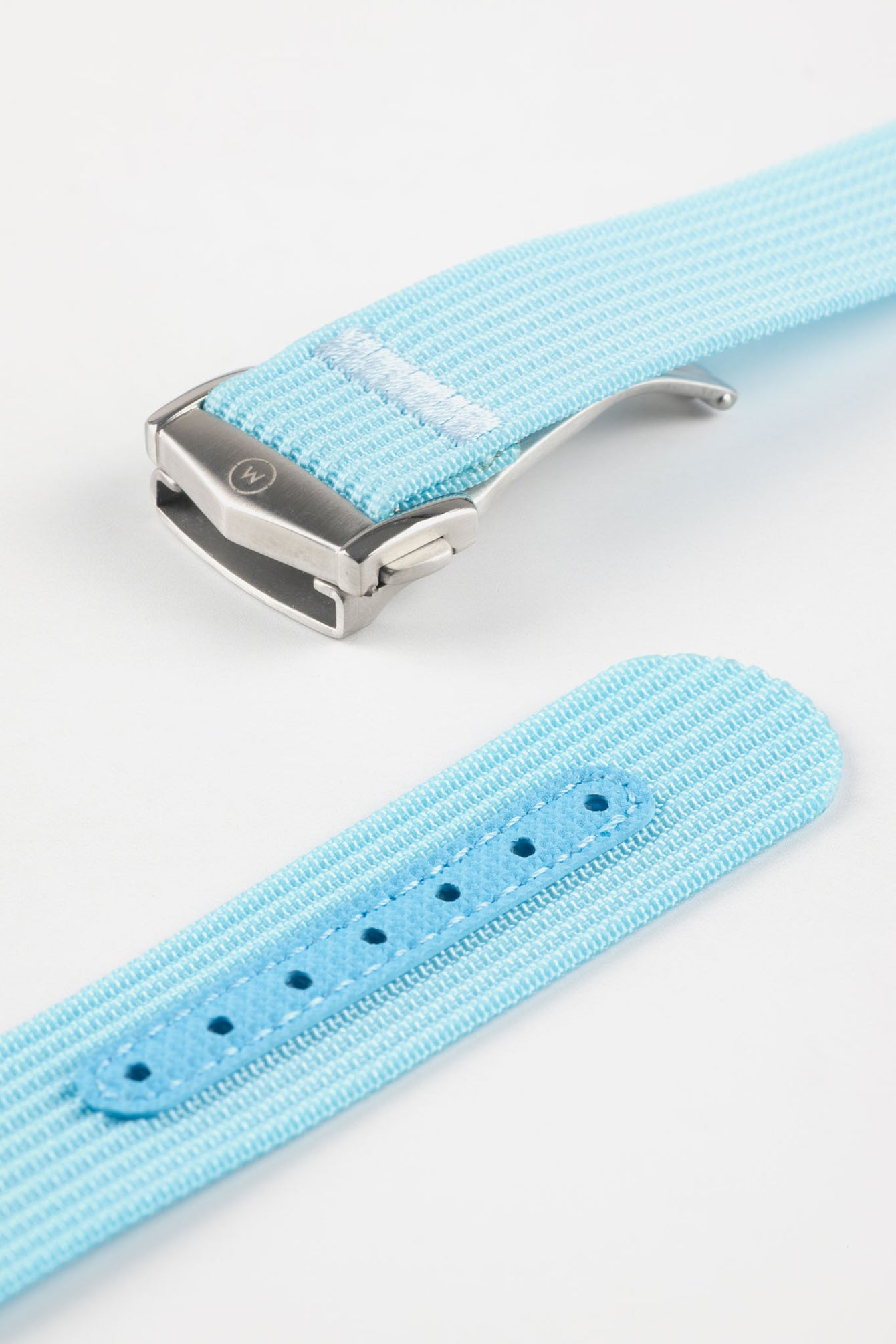 Angled close-up of a light turquoise ribbed nylon watch strap highlighting the fine woven texture and a brushed silver deployant clasp with an engraved logo. A separate strap section shows a matching turquoise reinforced panel with neatly stitched edges and evenly spaced adjustment holes, all displayed on a clean light grey background.