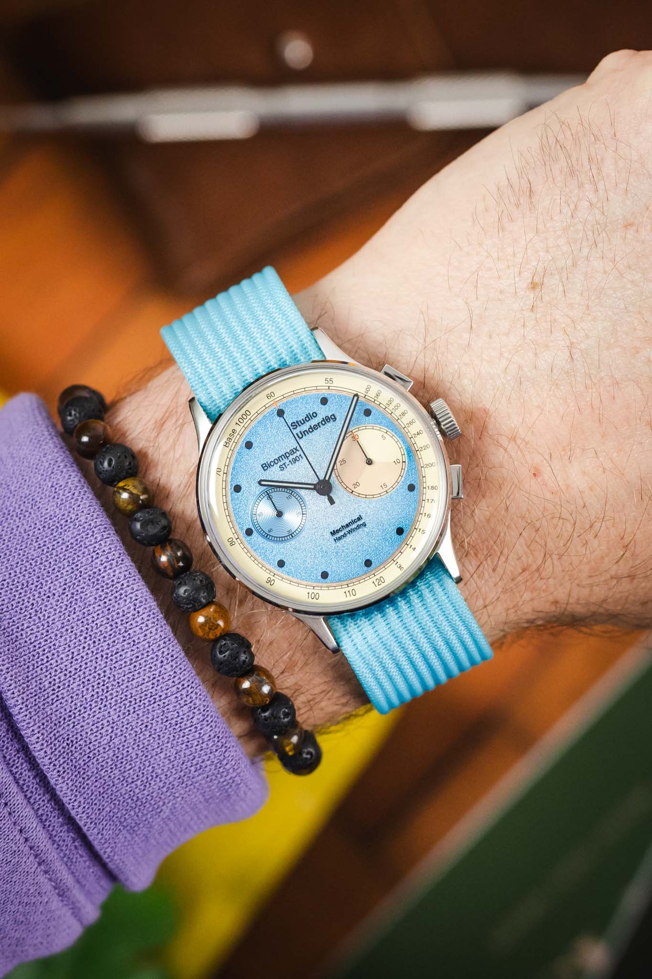 Close-up of a wrist wearing a silver chronograph watch with a textured light blue dial, cream subdials, and a turquoise ribbed fabric strap, paired with a dark beaded bracelet and a purple sleeve, set against a softly blurred warm-toned background.
