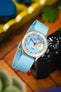 Close-up of a silver chronograph wristwatch with a textured light blue dial and turquoise ribbed fabric strap resting on a black camera body, with a blurred background featuring warm wood tones, a yellow tray, and green plant leaves.
