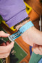Close-up of a wrist with a turquoise ribbed fabric watch strap being fastened with a metal clasp by hand, paired with a dark beaded bracelet and a purple sleeve, set against a colorful blurred background.