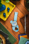 Flat lay of a silver chronograph wristwatch with a textured light blue dial and turquoise ribbed fabric strap placed on a wooden surface, surrounded by everyday items including blue wireless earbuds in a yellow tray, a leather pouch, a precision tool, magazines, and green plant leaves.