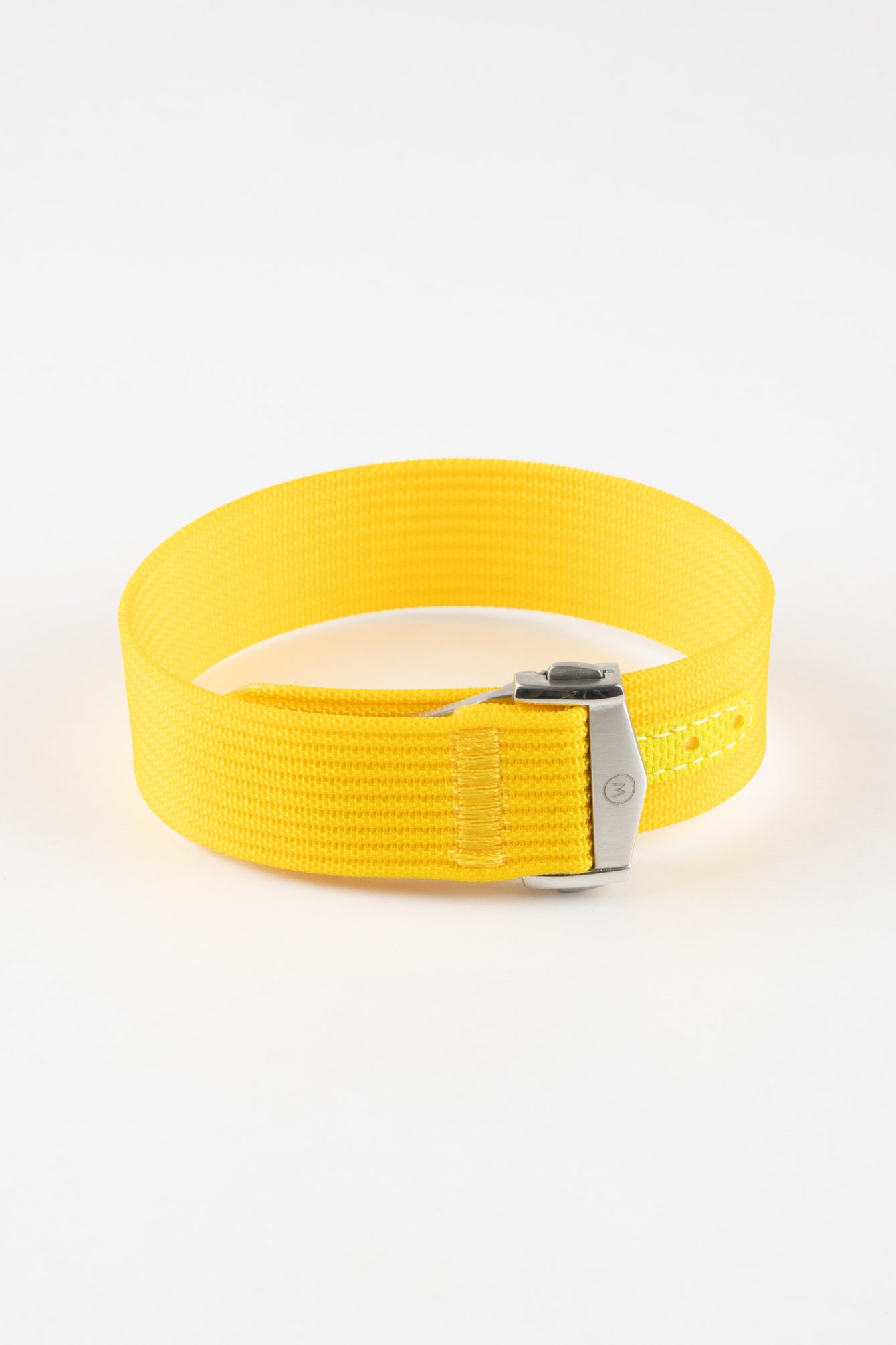Close-up of a bright yellow ribbed nylon watch strap arranged in a loop on a light grey background. The strap features a textured woven surface and a brushed silver deployant clasp with an engraved logo. A matching yellow reinforced section with neatly stitched edges and evenly spaced adjustment holes is visible near the clasp, emphasizing the strap’s construction and vibrant color.
