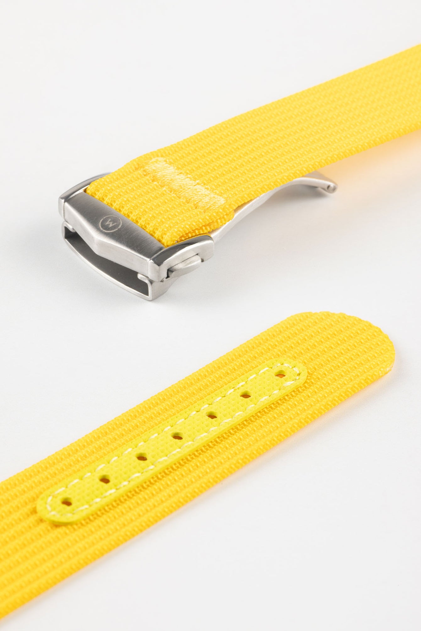 Angled close-up of a bright yellow ribbed nylon watch strap highlighting its textured woven surface and a brushed silver deployant clasp with an engraved logo. A separate strap section shows a matching yellow reinforced panel with clean stitching and evenly spaced adjustment holes, all set against a light grey background.